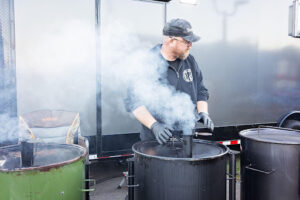 Pitmaster tending large barbecue smokers with steaming meat at an outdoor BBQ cooking setup, preparing smoked barbecue for an event or catering service.
